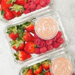 Three glass meal prep boxes with a round container of chocolate greek yogurt fruit dip surrounded by strawberries and raspberries