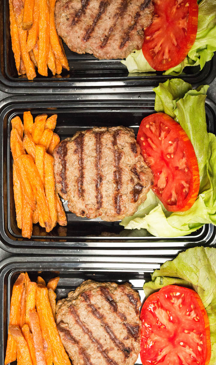 overhead shot of three black meal prep containers each with a hamburger patty, sweet potato fries, tomato, and lettuce