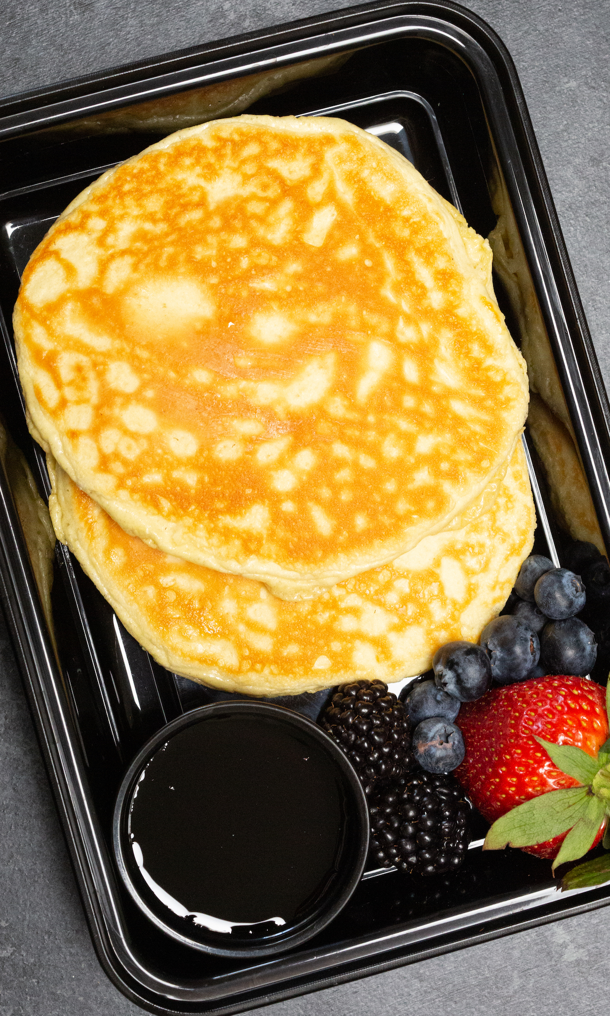 Overhead view of a single disposable meal prep container filled with two protein pancakes, syrup, blueberries, and strawberries.