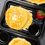 Overhead view of three black plastic meal prep containers each filled with two protein pancakes, strawberries & blueberries, and syrup