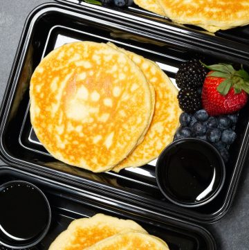 Overhead view of three black plastic meal prep containers each filled with two protein pancakes, strawberries & blueberries, and syrup