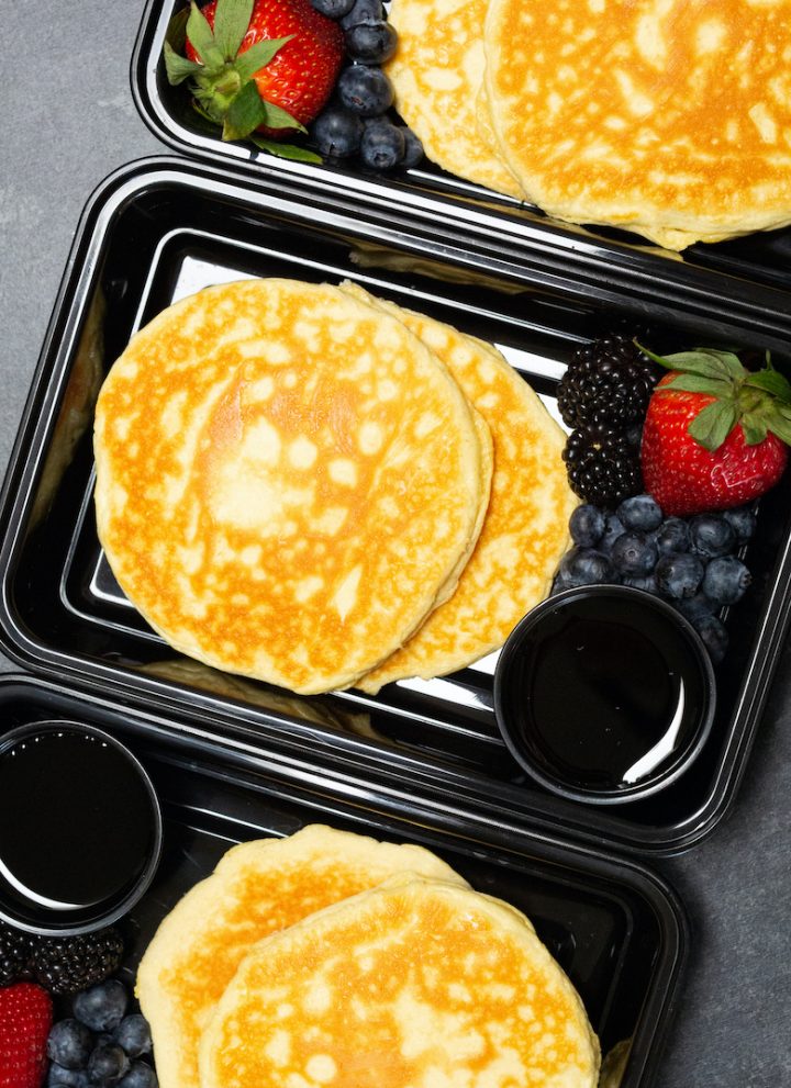 Overhead view of three black plastic meal prep containers each filled with two protein pancakes, strawberries & blueberries, and syrup