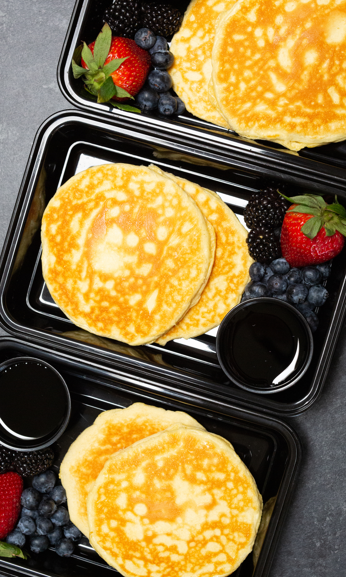 Overhead view of three black plastic meal prep containers each filled with two protein pancakes, strawberries & blueberries, and syrup