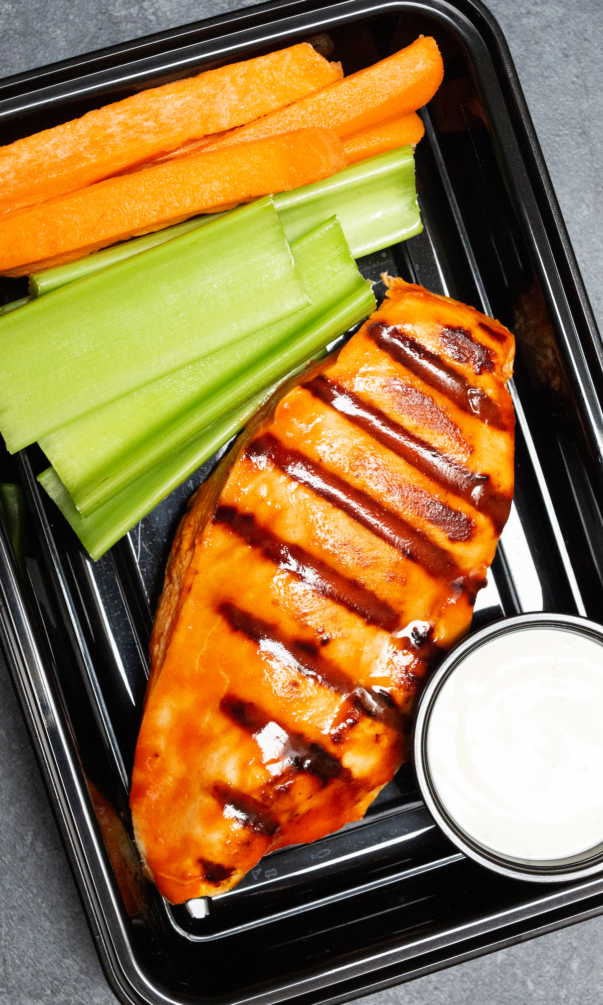 Overhead view of a disposable black plastic meal prep container that contains a grilled chicken breast slathered in orange buffalo sauce next to carrot and celery sticks and a small container of greek ranch.