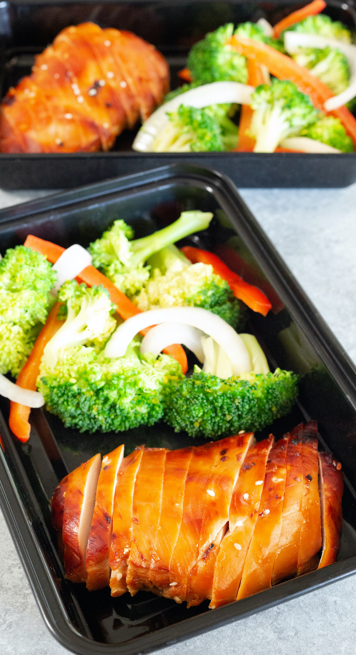 A black plastic meal prep container with a sliced teriyaki chicken breast and vegetables is in focus in the foreground, with a another container out of focus in the background.