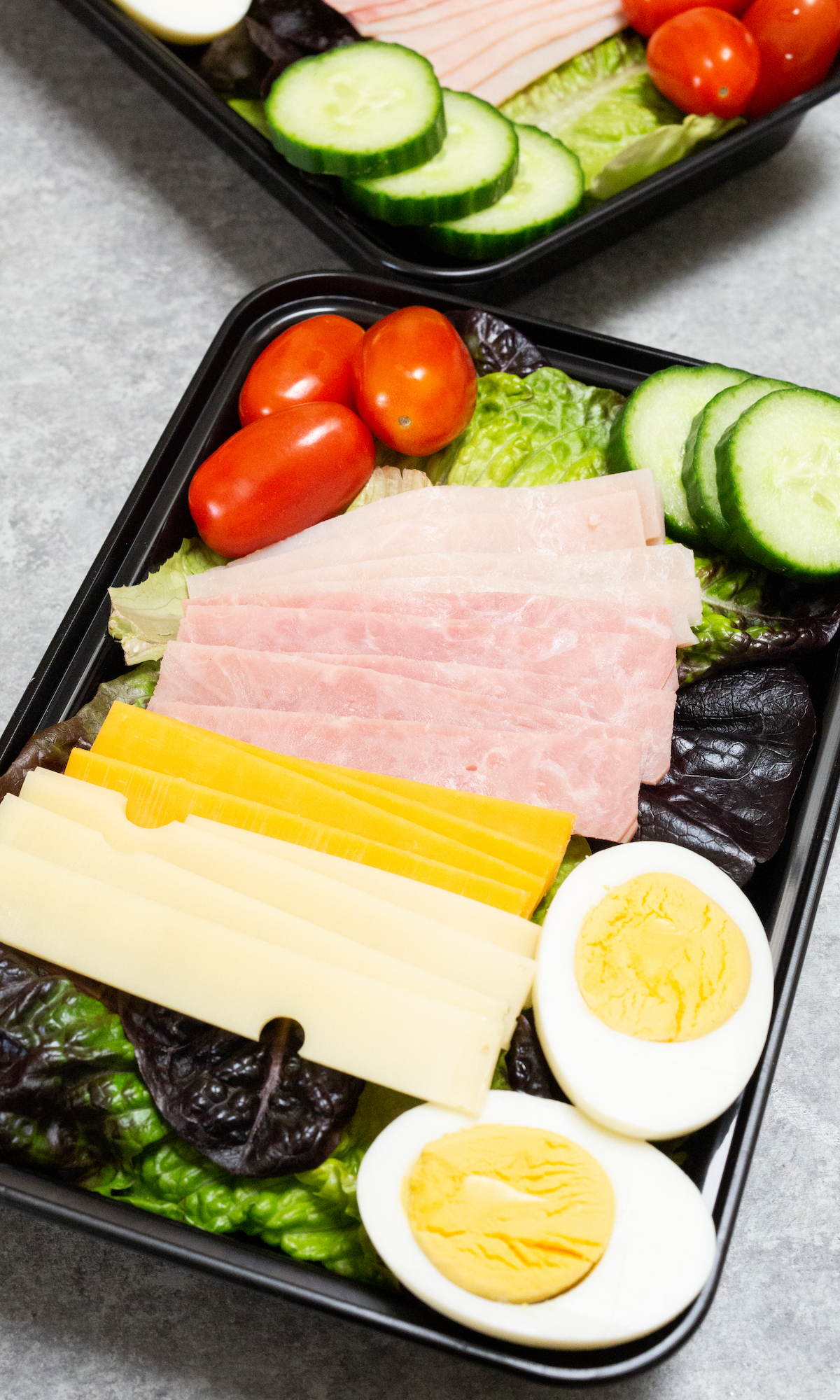 A black meal prep container is filled with greens, sliced deli meat, a hard boiled egg, cherry tomatoes, and sliced cucumbers.