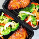 Overhead view of two black plastic meal prep containers filled with sliced teriyaki chicken breast and broccoli, peppers, and onions.