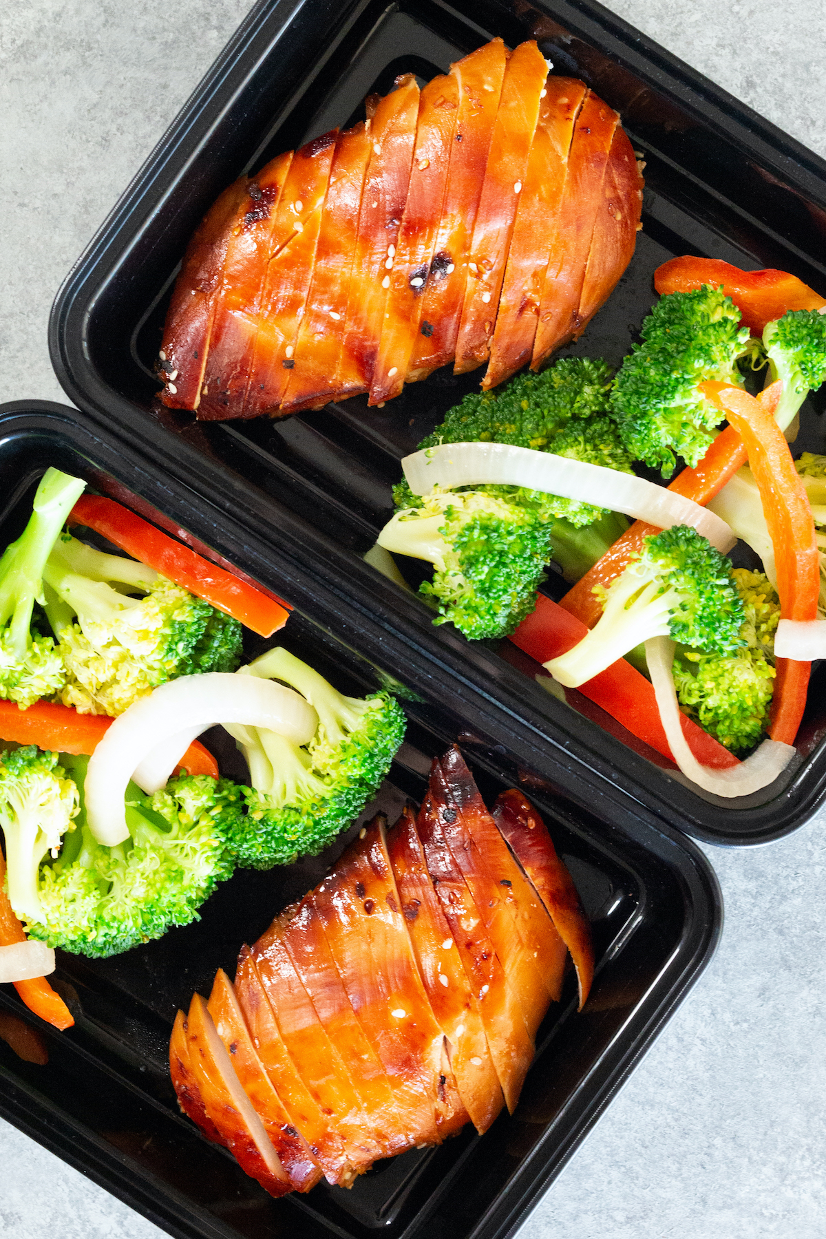 Overhead view of two black plastic meal prep containers filled with sliced teriyaki chicken breast and broccoli, peppers, and onions.