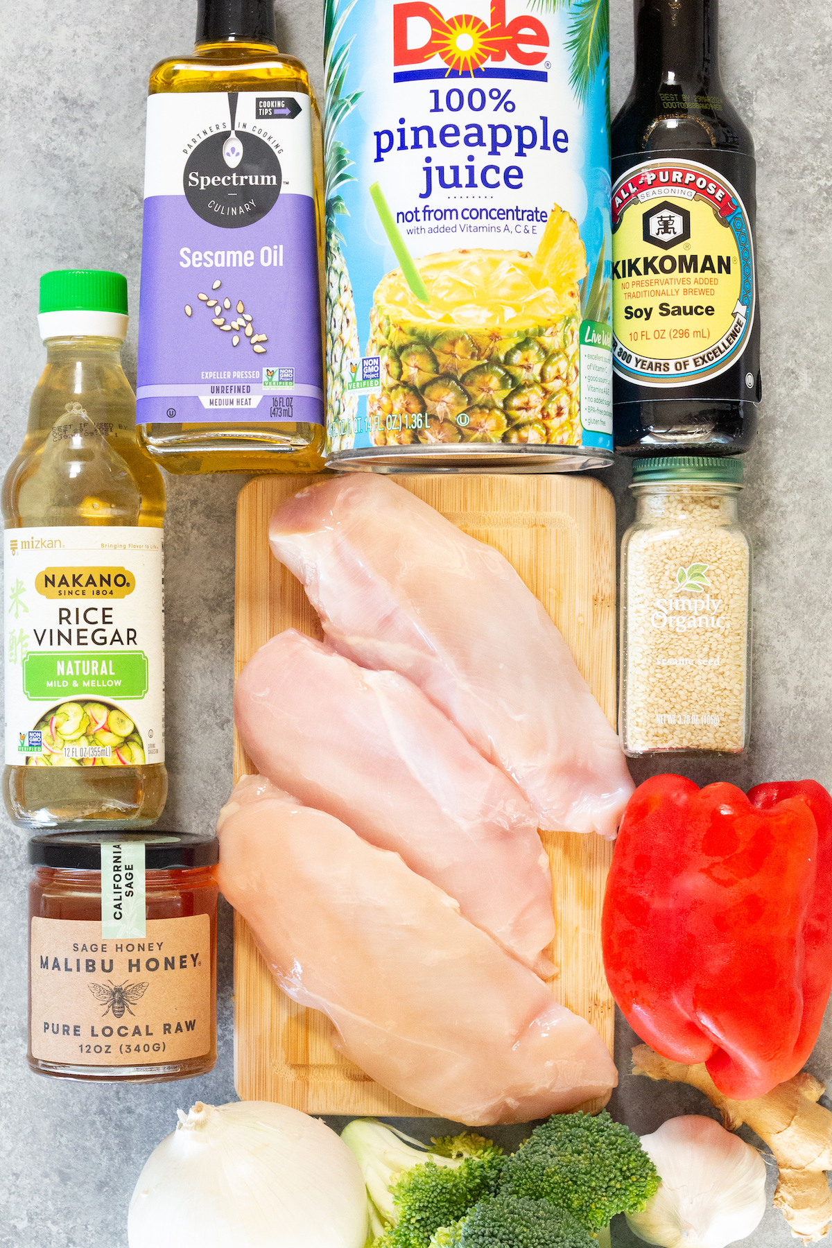 Overhead view of the ingredients to make teriyaki chicken meal prep. (Sesame oil, pineapple juice, soy sauce, rice vinegar, chicken breast, honey, sesame seeds, broccoli, onion, garlic, and ginger.)