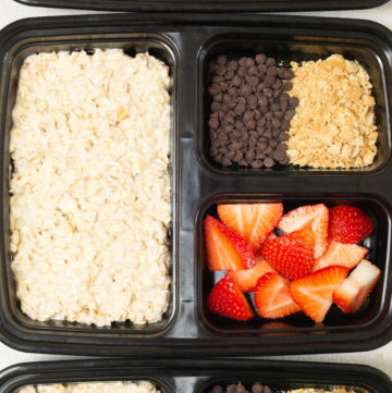 Overhead view looking down on. three black plastic divided meal prep containers. The large portion is filled with strawberry cheesecake overnight oats and the two smaller divided sections are filled with fresh sliced strawberries, crushed graham crackers, and mini chocolate chips.