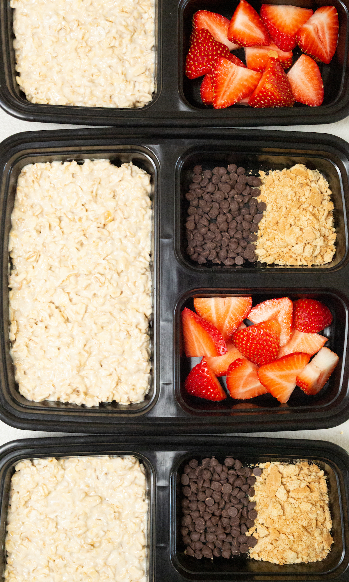 Overhead view looking down on three black plastic divided meal prep containers. The large portion is filled with strawberry cheesecake overnight oats and the two smaller divided sections are filled with fresh sliced strawberries, crushed graham crackers, and mini chocolate chips.