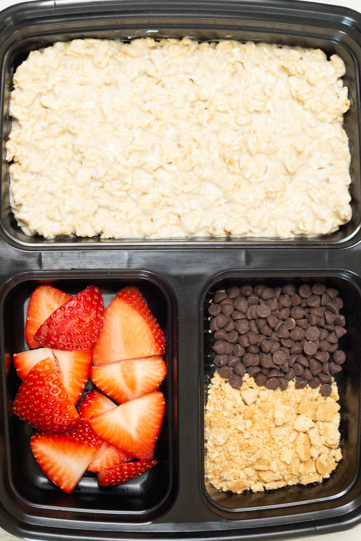 Close up of a single black plastic meal prep container that has three divided sections. The largest is filled with cheesecake overnight oats and the two smaller ones are filled with sliced strawberries, mini chocolate chips, and crushed graham crackers.