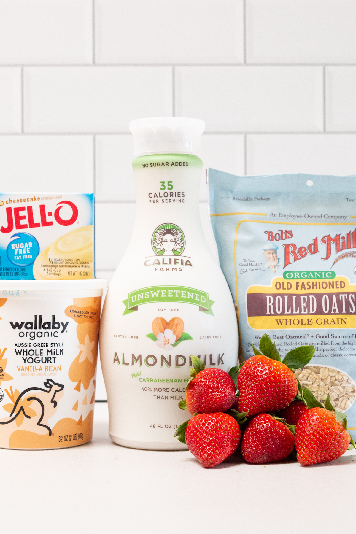 All the ingredients to make strawberry cheesecake overnight oats sit on a white background. A package of sugar-free Jello cheesecake pudding mix, vanilla bean Greek yogurt, unsweetened almond milk, fresh strawberries, and rolled oats.