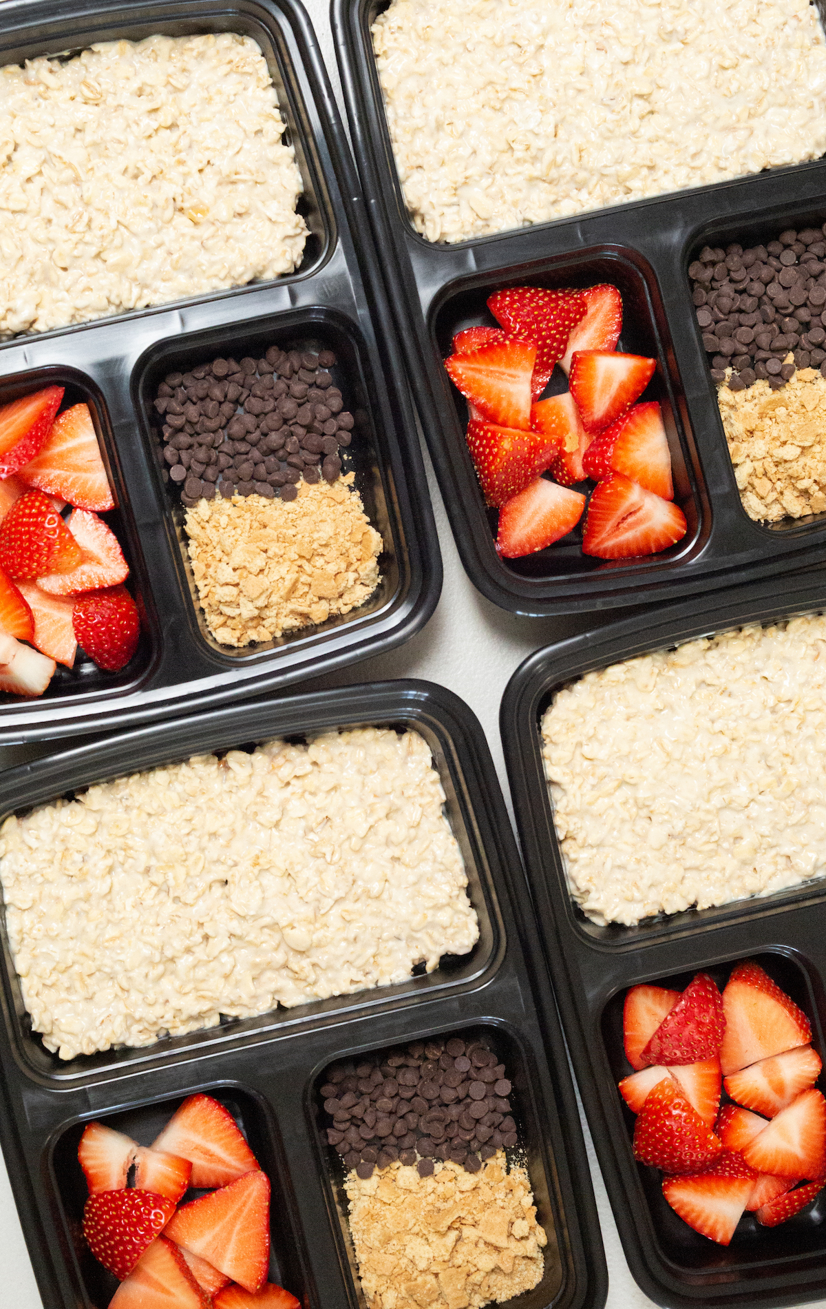 Overhead look at four black divided meal prep containers filled with cheesecake overnight oats, sliced strawberries, crushed graham crackers, and mini chocolate chip.s