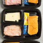 Overhead view of three plastic 3-compartment meal prep containers, filled with bread, sliced cheese, sliced meat, and condiment packets.