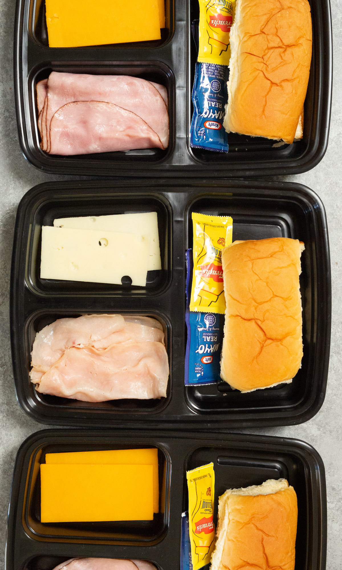 Overhead view of three plastic 3-compartment meal prep containers, filled with bread, sliced cheese, sliced meat, and condiment packets.