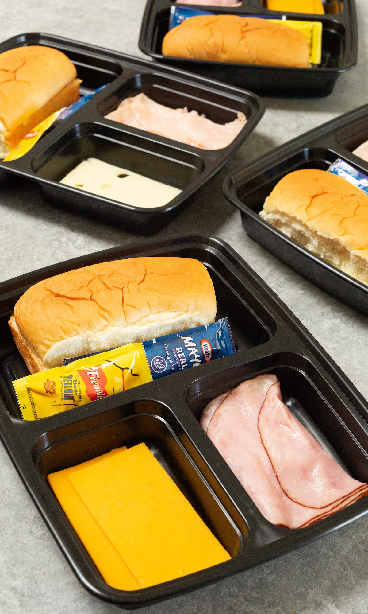 Four three compartment black plastic meal prep containers on a light background. Each individual compartment is filled with bread, sliced cheese, or sliced meat and condiment packets.