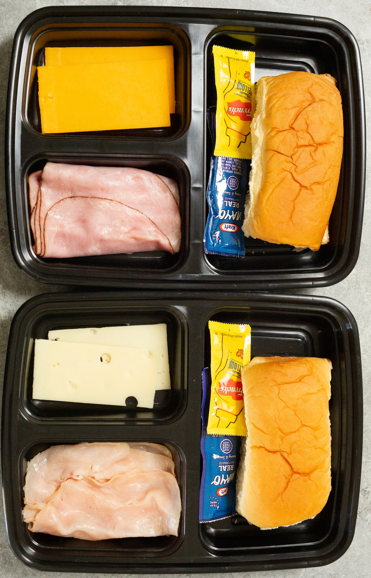 Overhead view of two black plastic meal prep containers filled with the ingredients to make a meat and cheese sandwich.