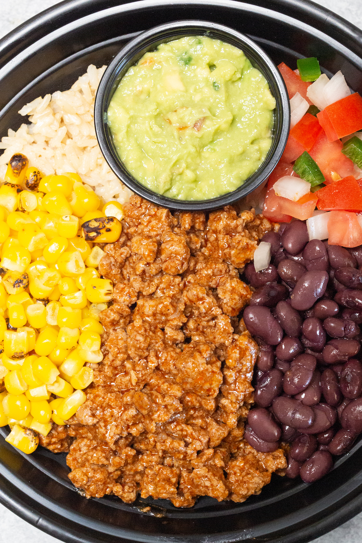 Close up of the ingredients in a taco bowl meal prep featuring taco seasoned ground beef, beans, corn, rice, salsa, and guacamole.