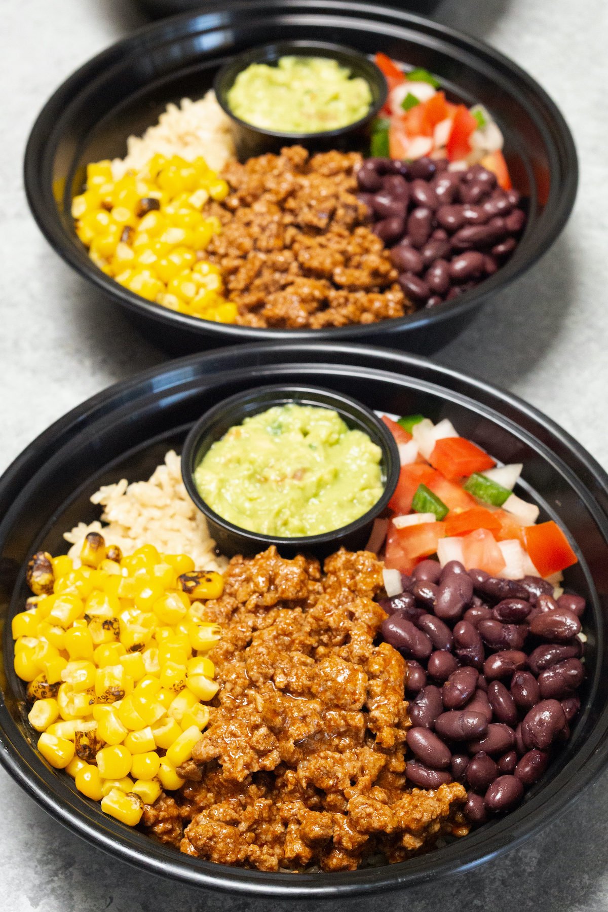 Two plastic meal prep bowls filled with taco meat, beans, corn, salsa, rice, and guacamole.