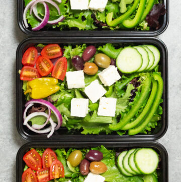 Overhead look at three black plastic meal prep containers filled with Greek salads that are topped with feta cubes, sliced cherry tomatoes, sliced red onion, cucumber, and bell pepper.