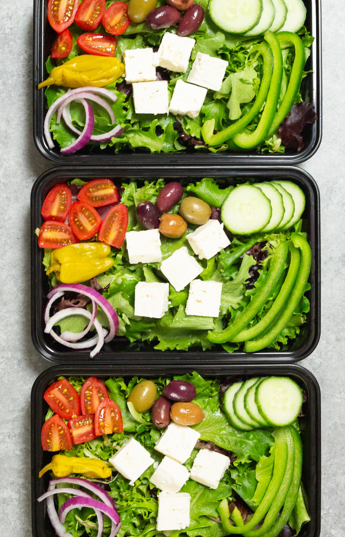 Overhead look at three black plastic meal prep containers filled with Greek salads that are topped with feta cubes, sliced cherry tomatoes, sliced red onion, cucumber, and bell pepper.