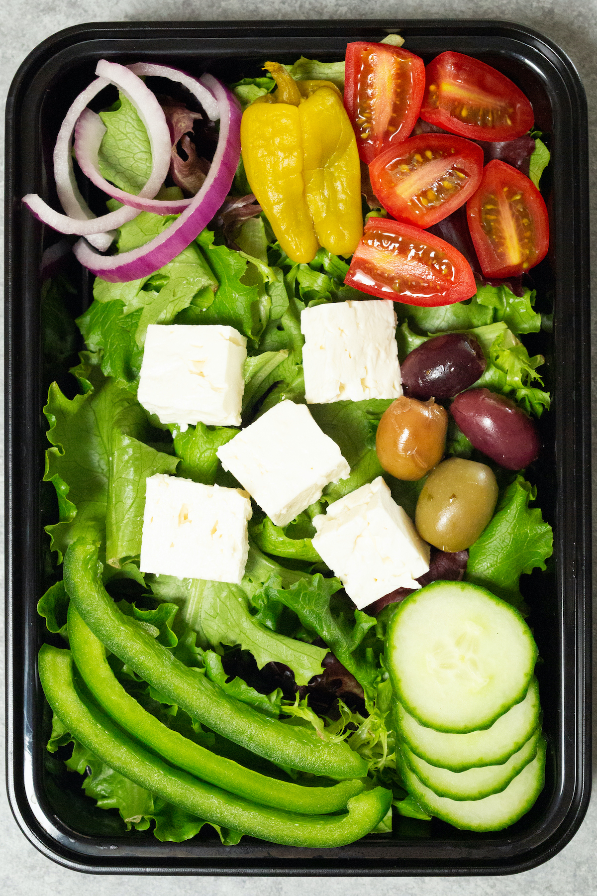 A single black plastic meal prep container filled with a Greek salad that's topped with cubed feta, sliced bell peppers, cucumber, tomatoes, and red onion.