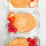 Three glass meal prep containers each filled with two chocolate protein pancakes and sliced strawberries and raspberries.