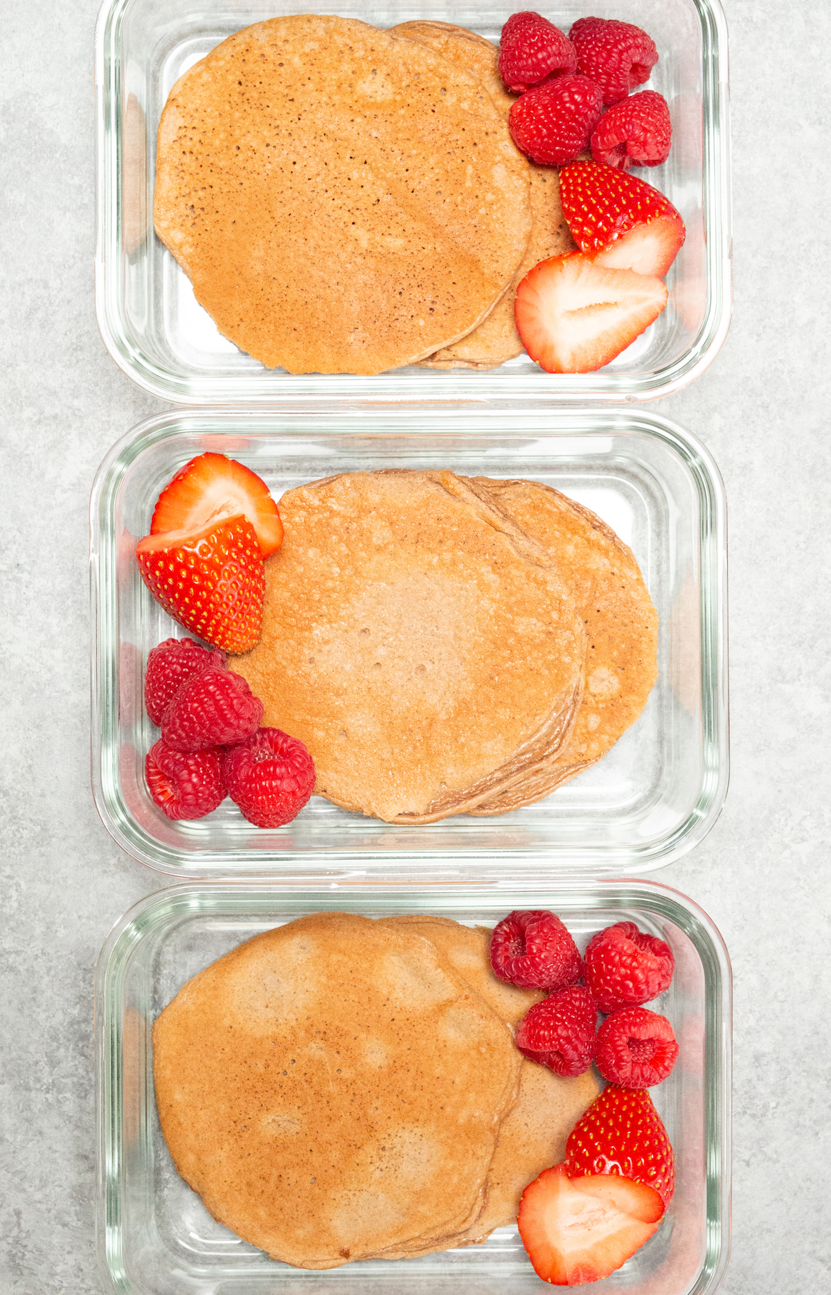 Three glass meal prep containers each filled with two chocolate protein pancakes and sliced strawberries and raspberries.