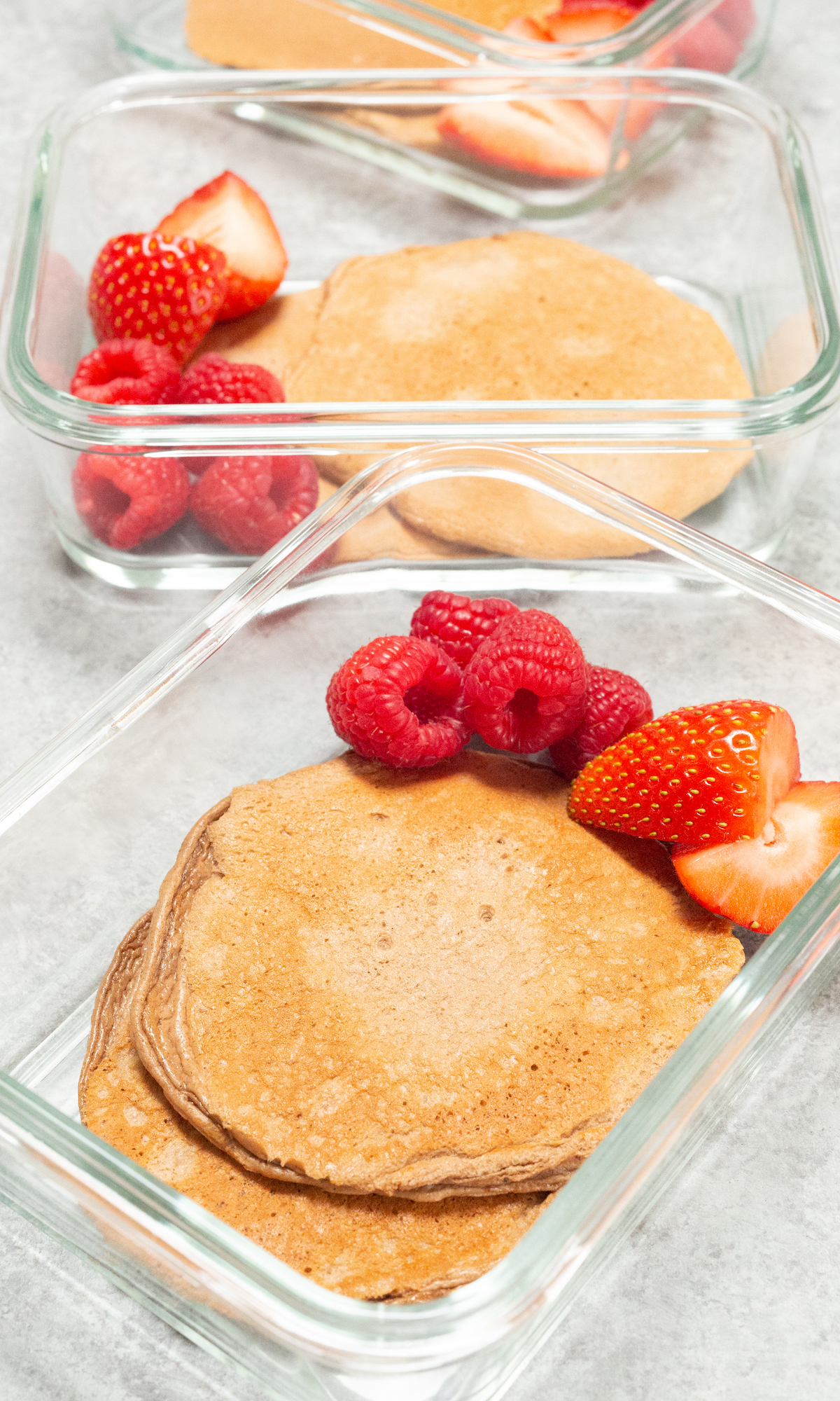 Glass meal prep containers filled with chocolate protein pancakes and red berries on a white background.