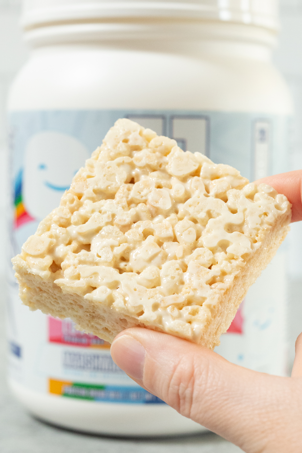 A hand holds hope a single protein rice krispy square in front of a tub of marshmallow protein powder.