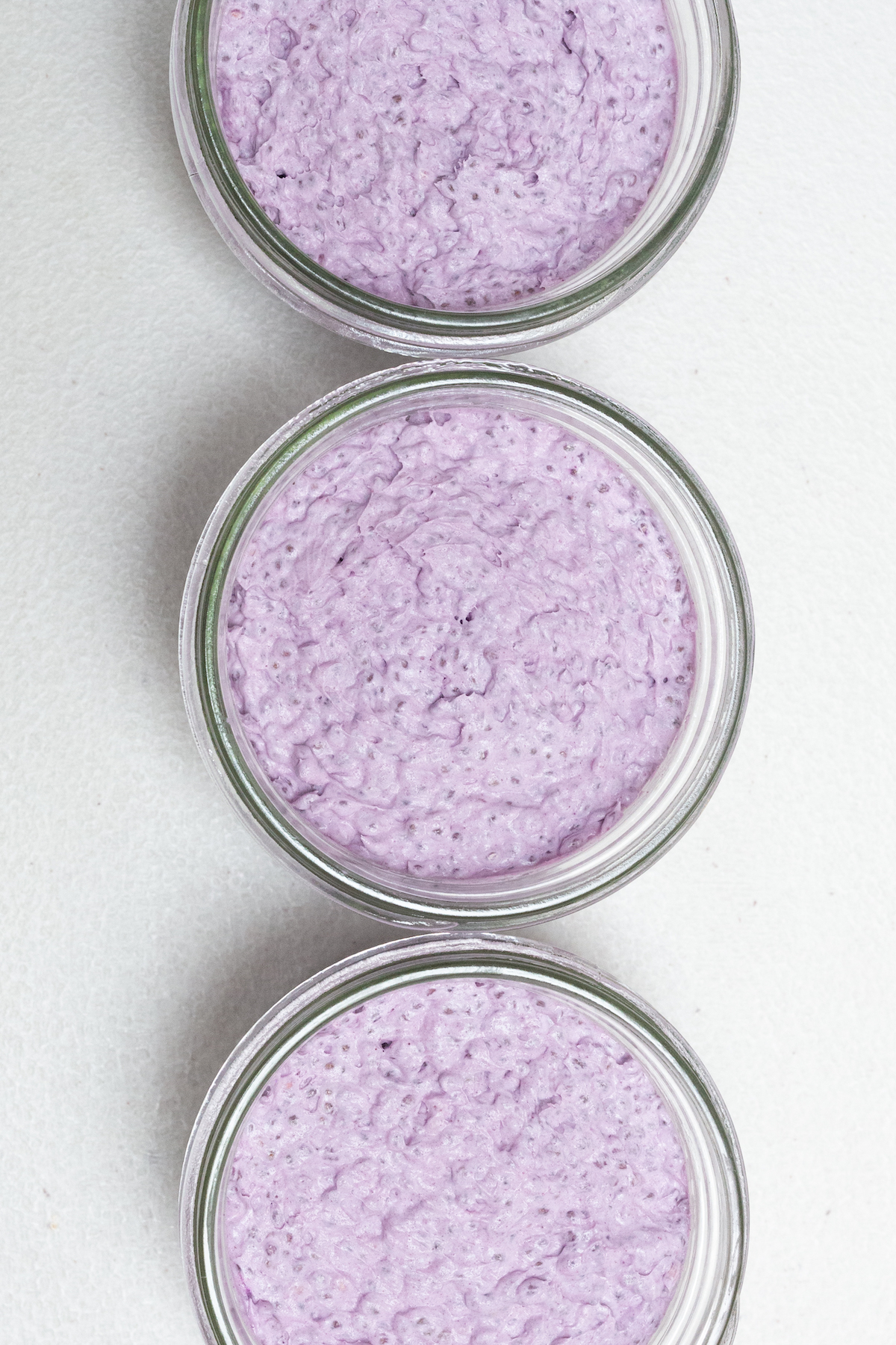 Overhead view of three small mason jars are filled with a thick purple colored ube chia pudding on a white background.