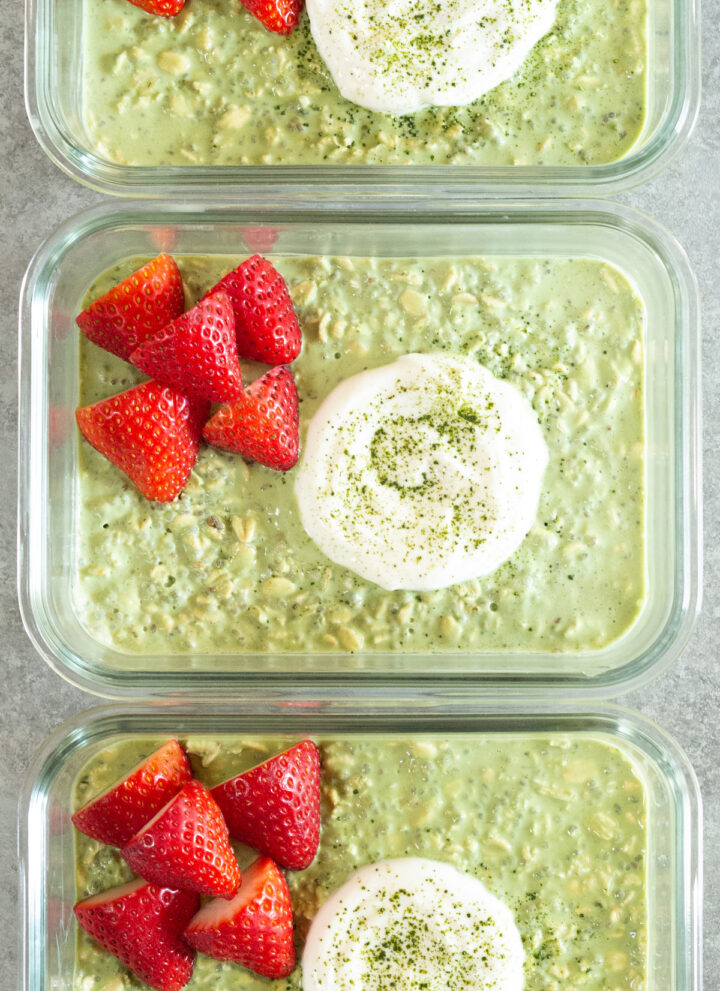 Three glass meal prep containers are filled with green matcha overnight oats. Each is topped with a dollop of Greek yogurt and several slices of strawberry.
