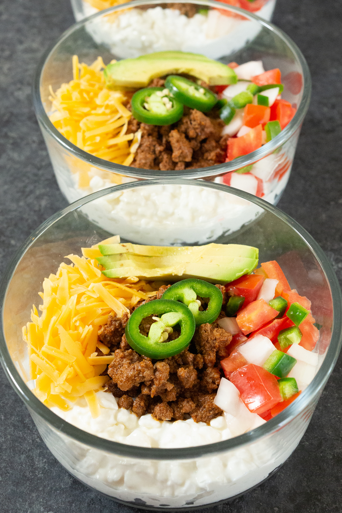 Two glass single serving pyrex bowls filled with cottage cheese, cooked ground beef, diced tomato, diced onion, diced and sliced jalapeno, shredded cheddar cheese, and sliced avocado.