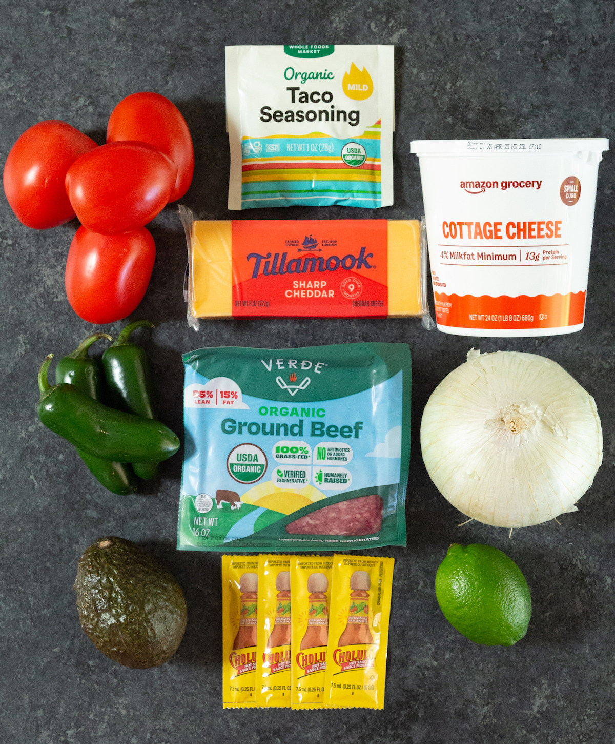 A packet of taco seasoning, four roma tomatoes, three jalapenos, an avocado, a white onion, a lime, 1 lb ground beed, a block of sharp cheddar, a large container of cream cheese, and four single serve Cholula packets.