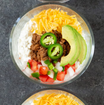 Three glass bowls are filled with cottage cheese, cooked ground beef, pico de gallo, shredded cheddar cheese, and avocado slices.