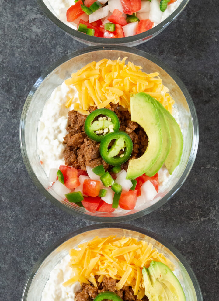 Three glass bowls are filled with cottage cheese, cooked ground beef, pico de gallo, shredded cheddar cheese, and avocado slices.