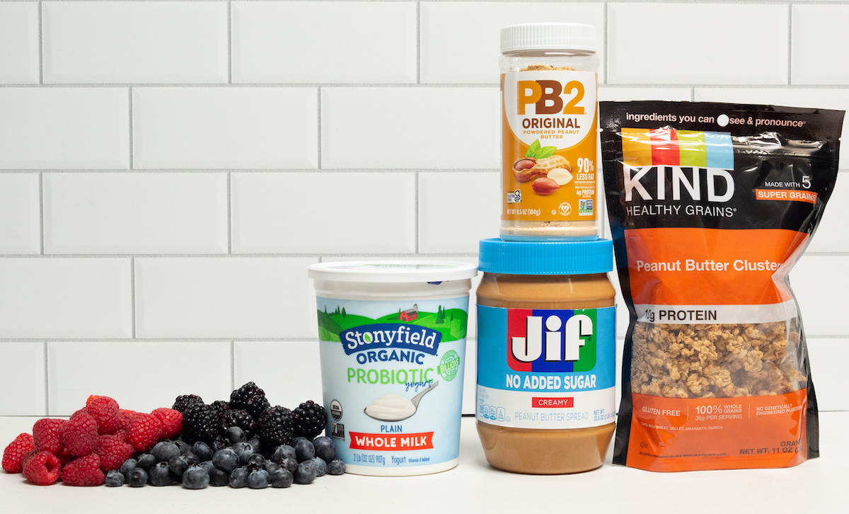 A medley of raspberries, blueberries, and blackberries sit next to a yogurt container, a peanut butter jar, a PB2 jar, and a bag of peanut butter granola clusters.