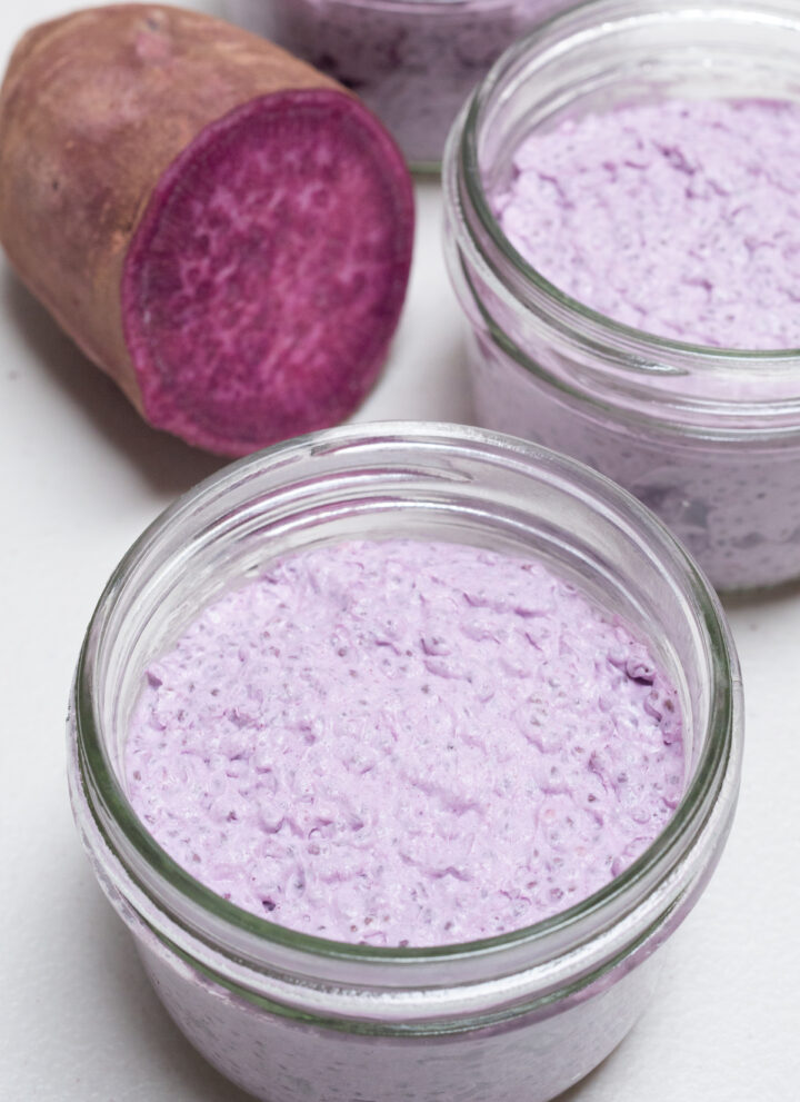A small mason jar filled with a light purple colored ube chia pudding is in focus in the foreground. More jars of the pudding and a purple sweet potato that's been cut in half are out of focus in the background.