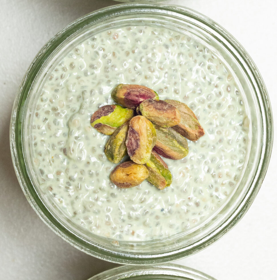 Three small mason jars filled with green pistachio chia pudding. A pile of shelled pistachios sits on top of the chia pudding.