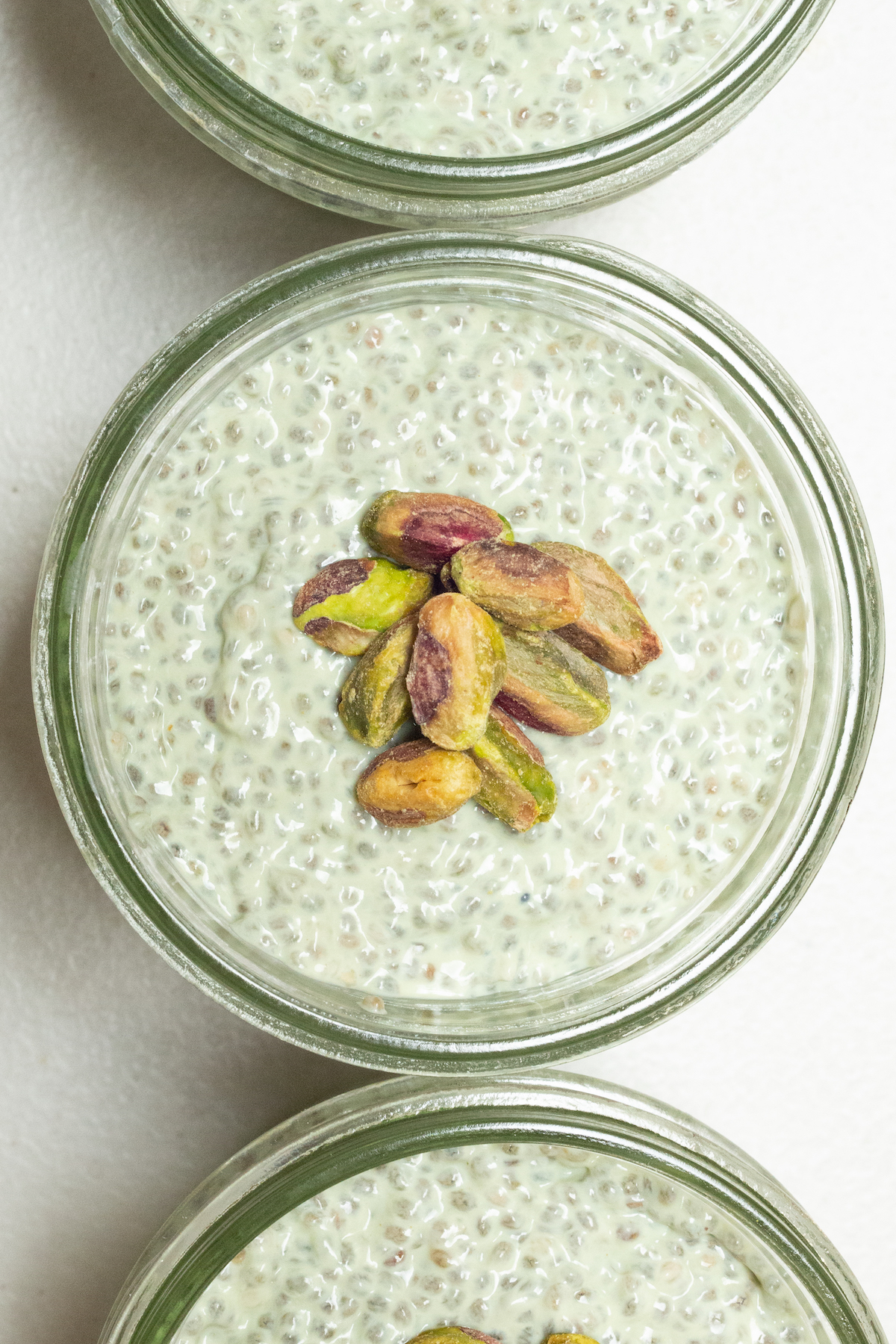 Three small mason jars filled with green pistachio chia pudding. A pile of shelled pistachios sits on top of the chia pudding.