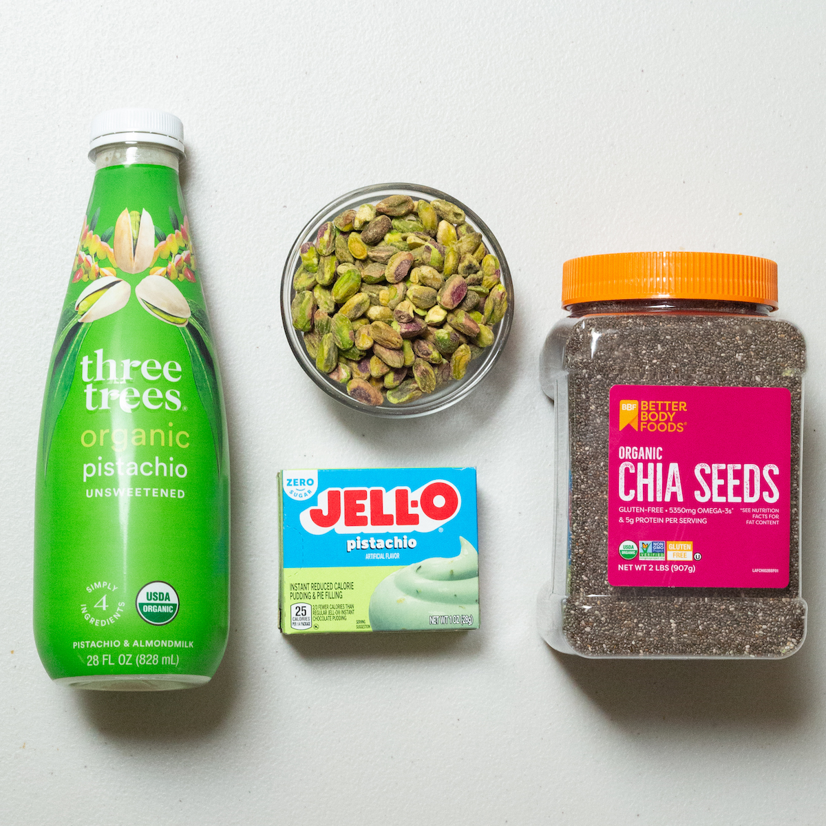 A bottle of pistachio milk, a box of sugar-free pistachio Jello pudding mix, shelled pistachios, and a container of chia seeds.