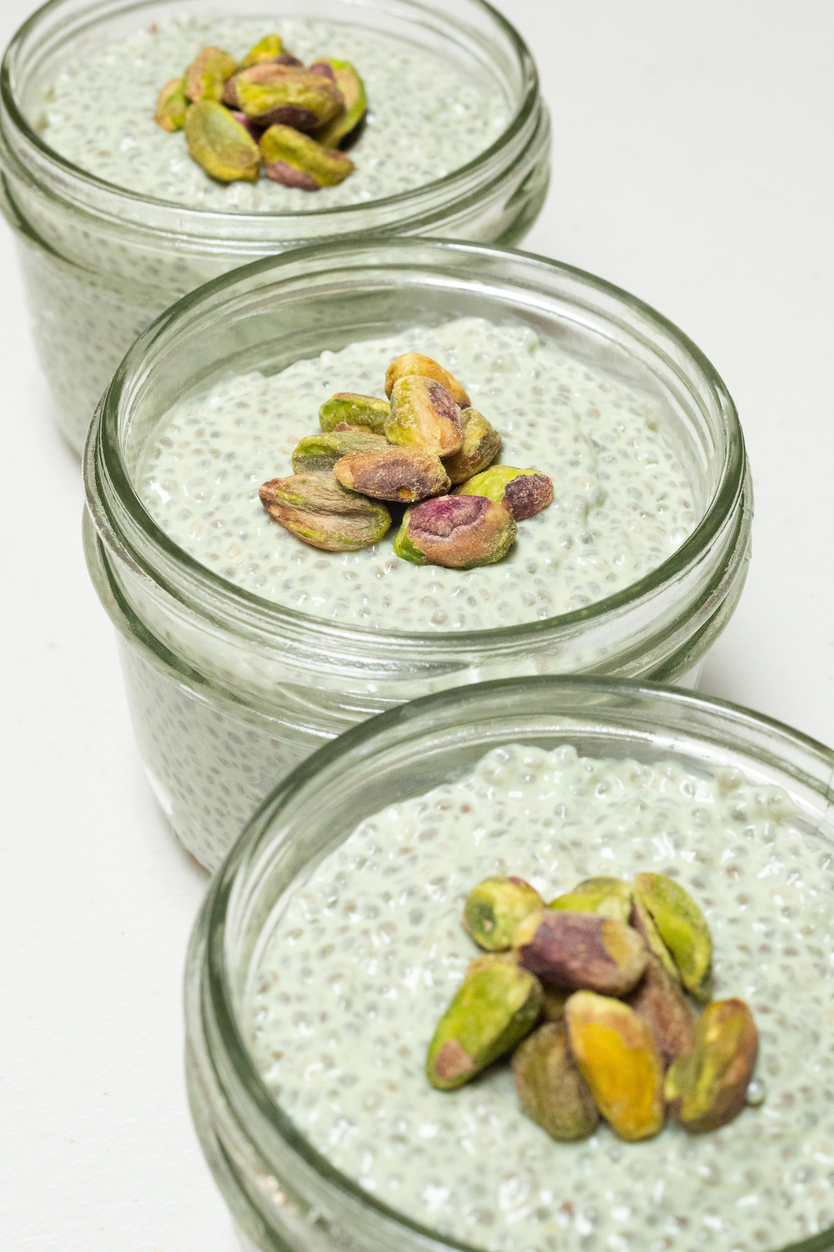 Three small mason jars filled with green pistachio chia pudding that's topped with shelled pistachios.