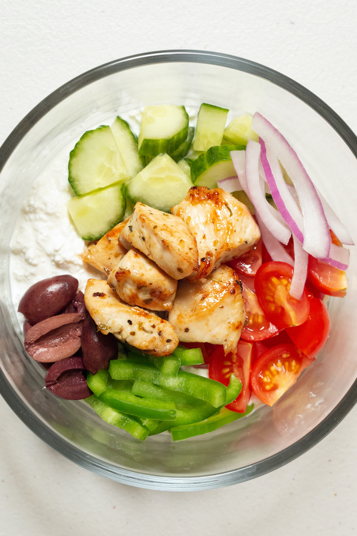 A glass bowl filled with cottage cheese that's topped with chopped green bell pepper, cherry tomatoes, red onion, cucumbers, kalamata olives, and cubed chicken breast.