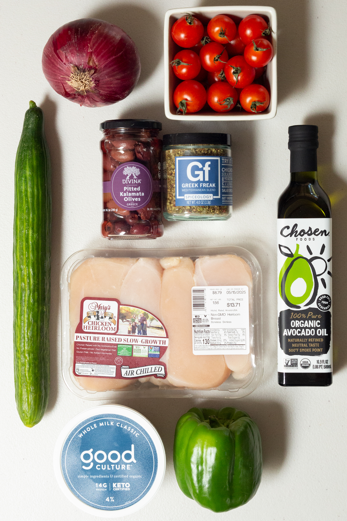 An english cucumber, a red onion, a pint of cherry tomatoes, a bottle of avocado oil, Greek seasoning, Kalamata olives, raw chicken breast, a tub of cottage cheese, and a green bell pepper on a white background.
