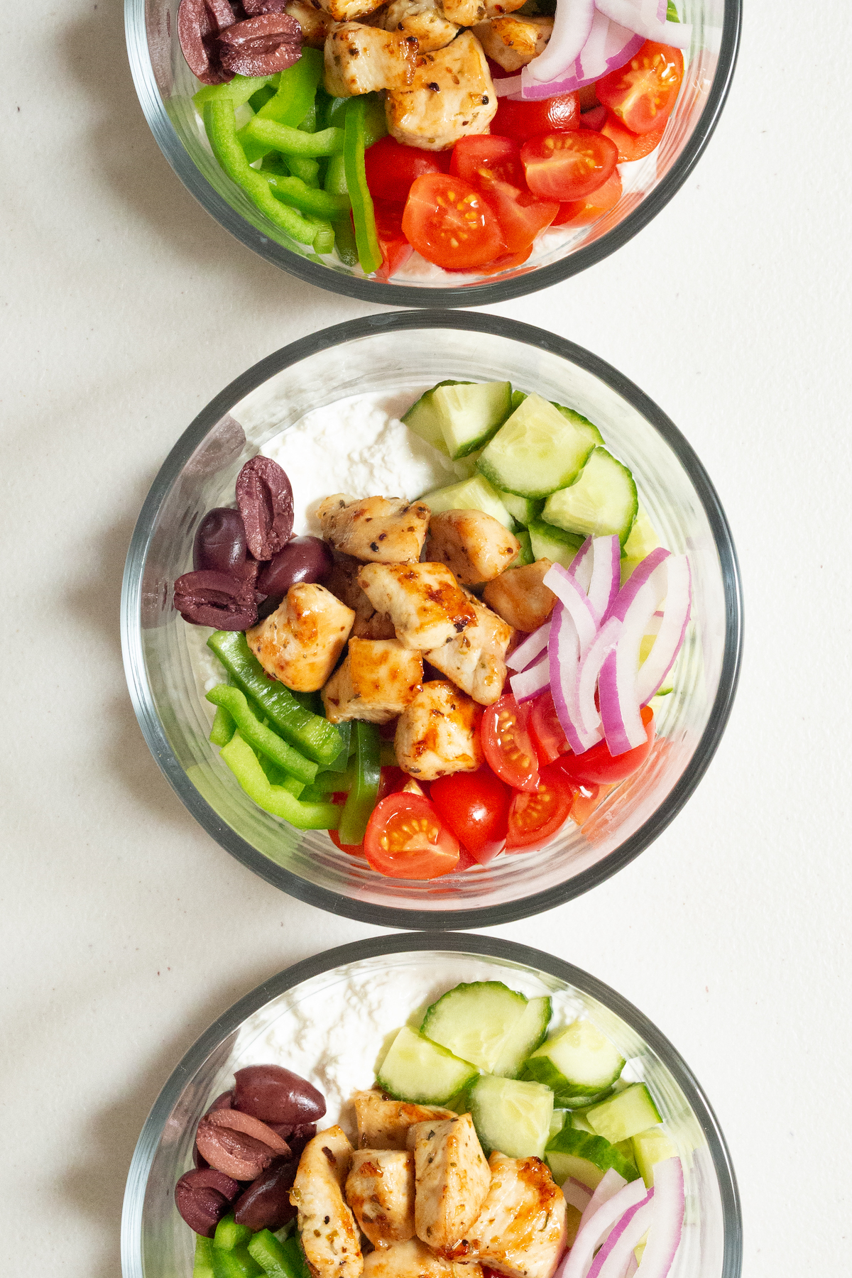Three glass pyrex bowls filled with cottage cheese topped with cubed, seasoned chicken, bell pepper strips, halved cherry tomatoes, diced cucumber, sliced red onion, and halved kalamata olives.
