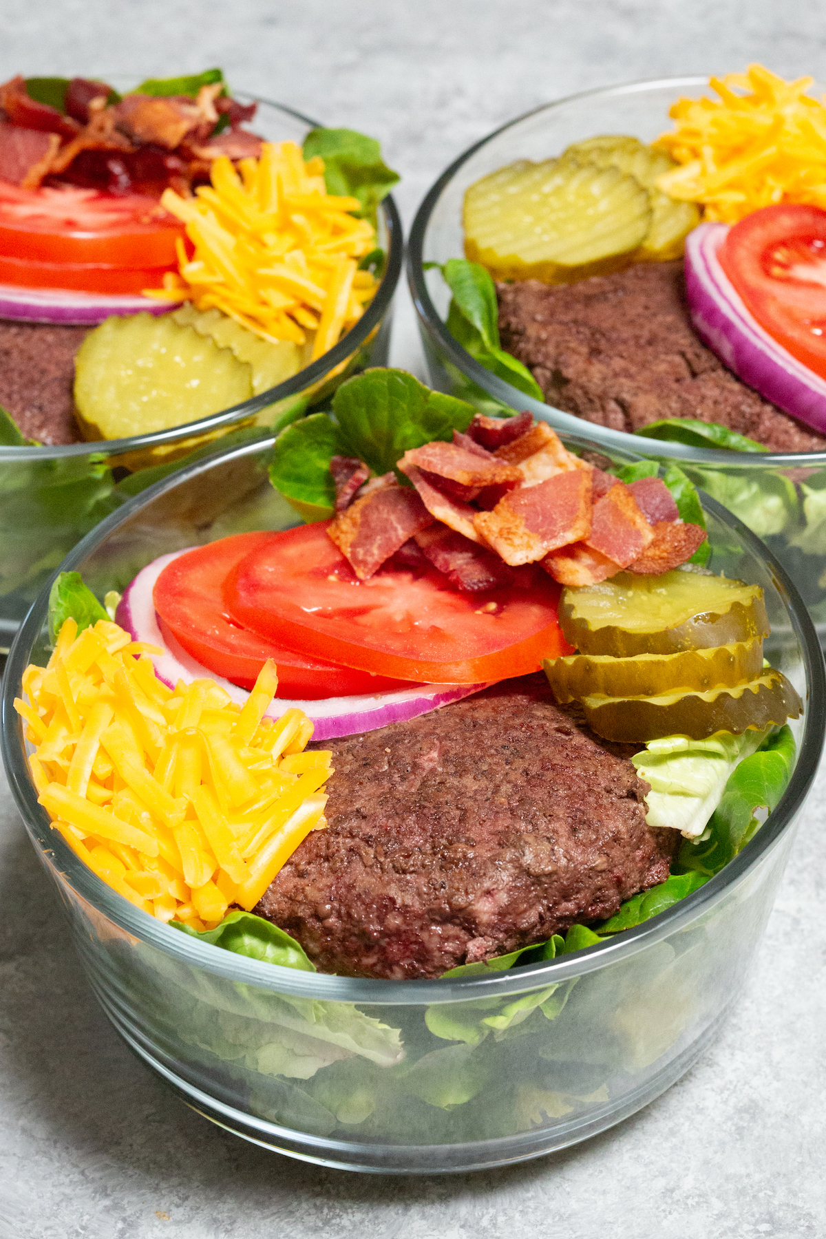 A glass bowl is in focus in the foreground. It is filled with butter crunch lettuce, a whole cooked burger patty, shredded yellow cheese, dill pickle chips, tomato & red onion slices, and chopped bacon. Two more bowls are out of focus in the background.