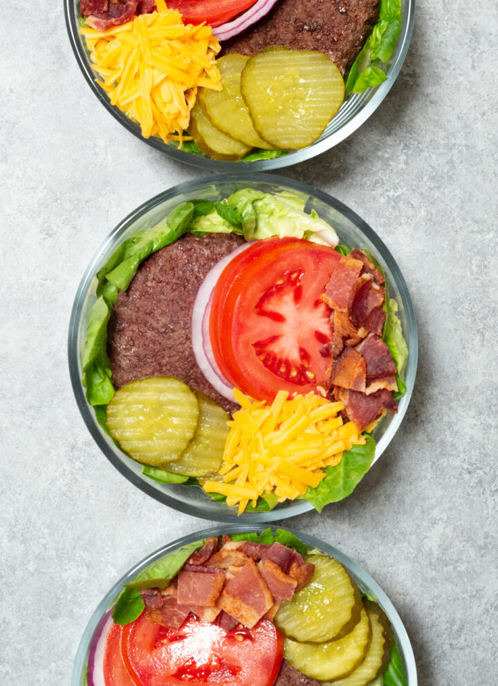 Three glass bowls filled with a bed of green lettuce that's topped with a cooked burger patty, sliced tomatoes, sliced red onions, pickled, bacon, and shredded cheddar cheese.