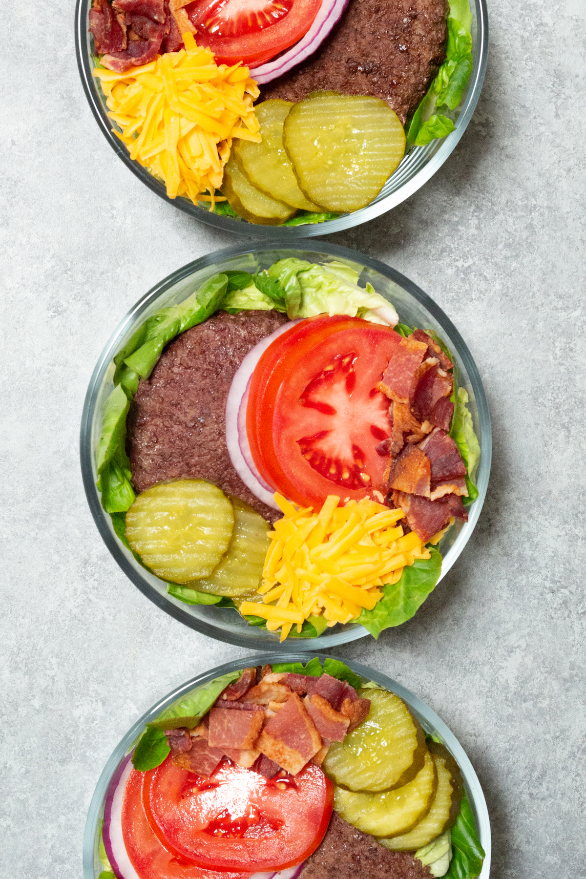 Three glass bowls filled with a bed of green lettuce that's topped with a cooked burger patty, sliced tomatoes, sliced red onions, pickled, bacon, and shredded cheddar cheese.
