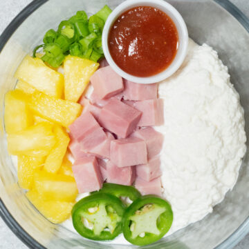 A clear glass bowl is filled with cottage cheese, ham chunks, pineapple chunks, sliced green onions, and sliced jalapenos.
